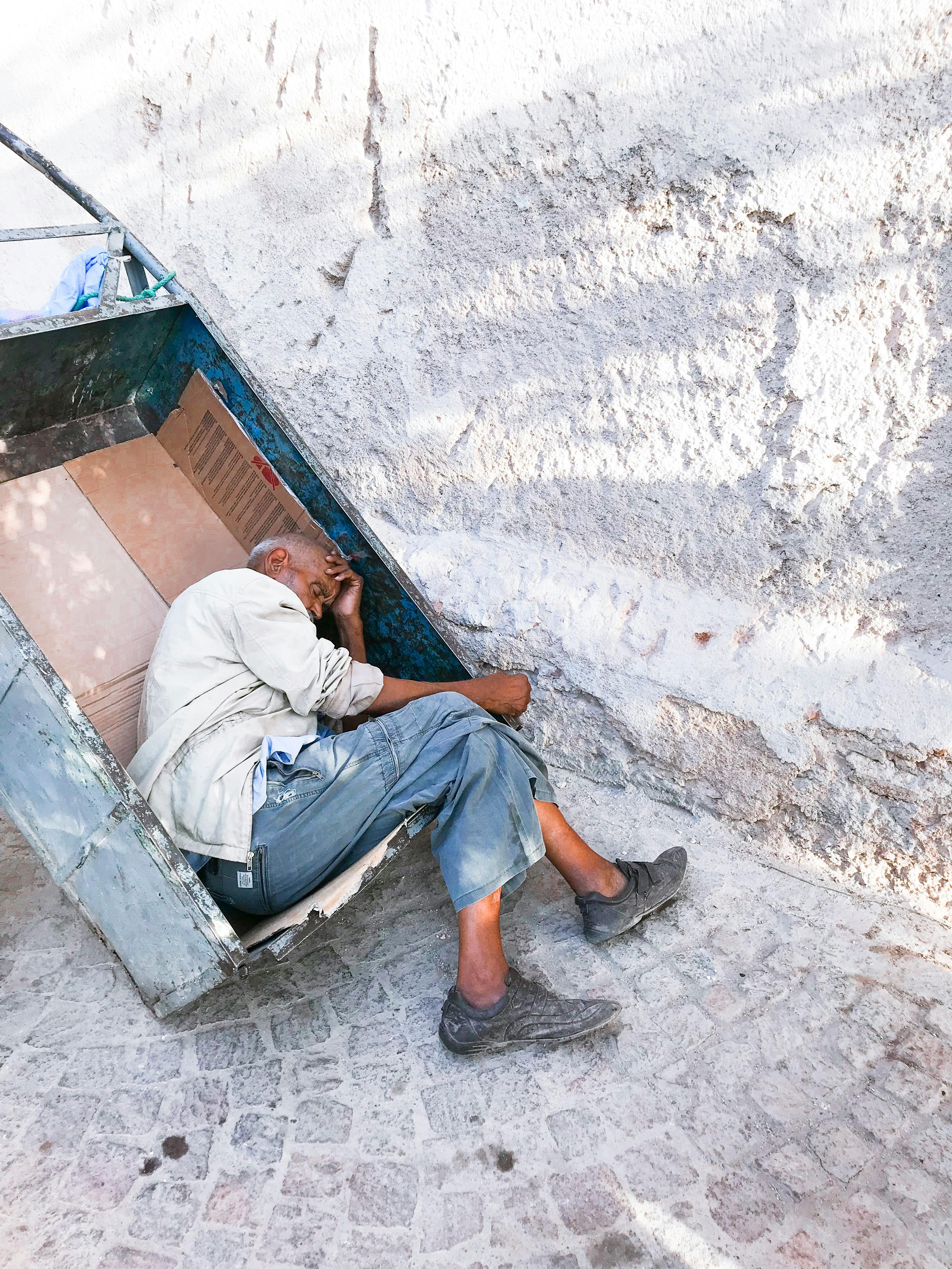 an elderly man sleeping on a metal cart on the of a street close to a wall