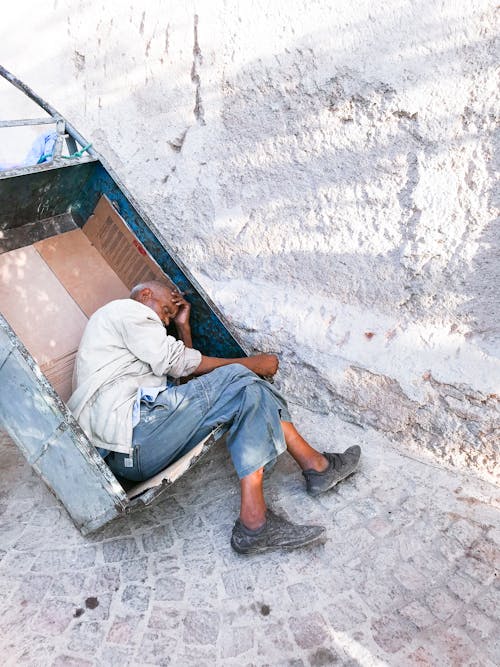 An Elderly Man Sleeping On A Metal Cart On The Of A Street Close To A Wall