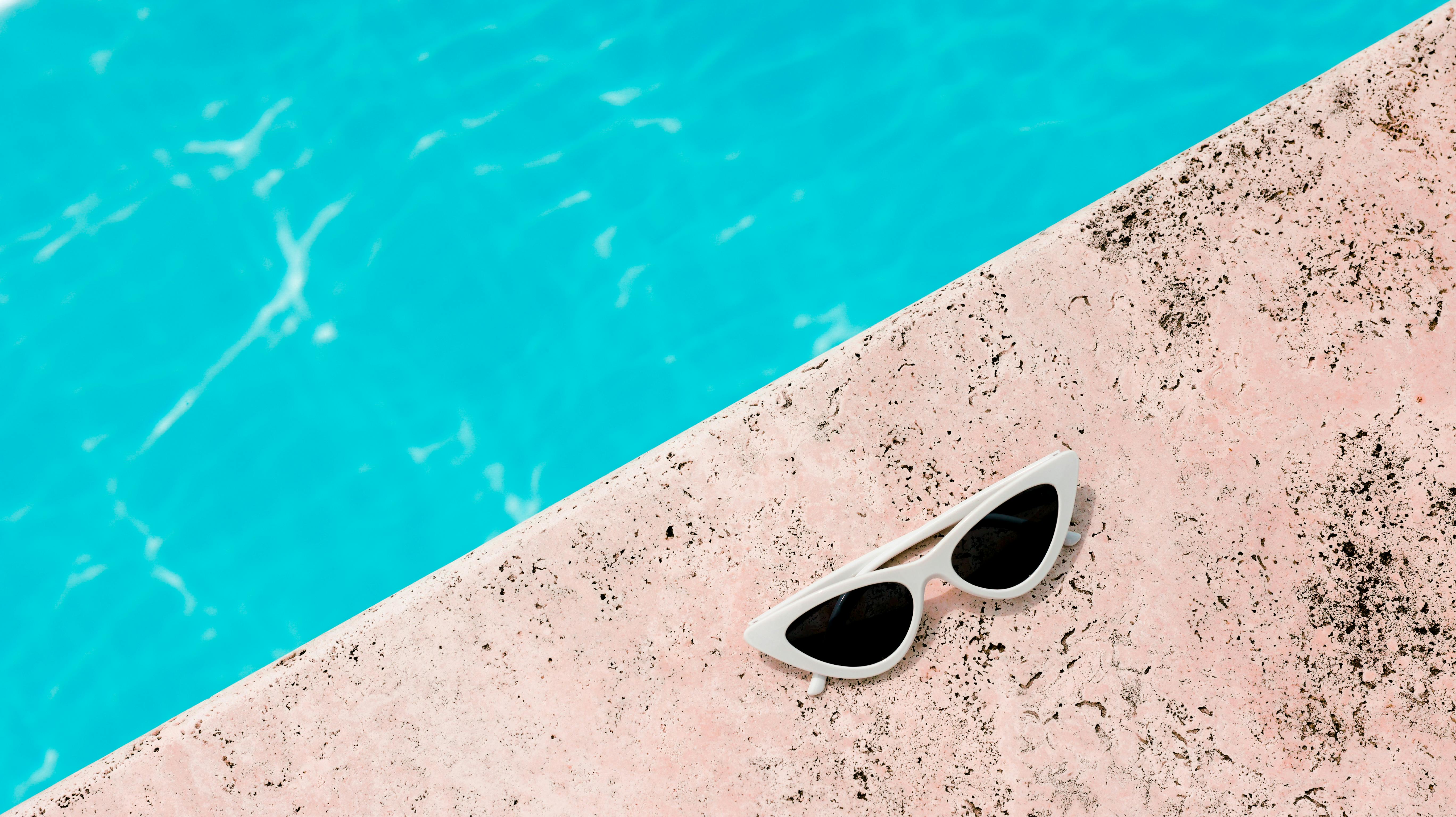 Chic white sunglasses resting on a pink poolside next to clear blue water, perfect for summer vibes.
