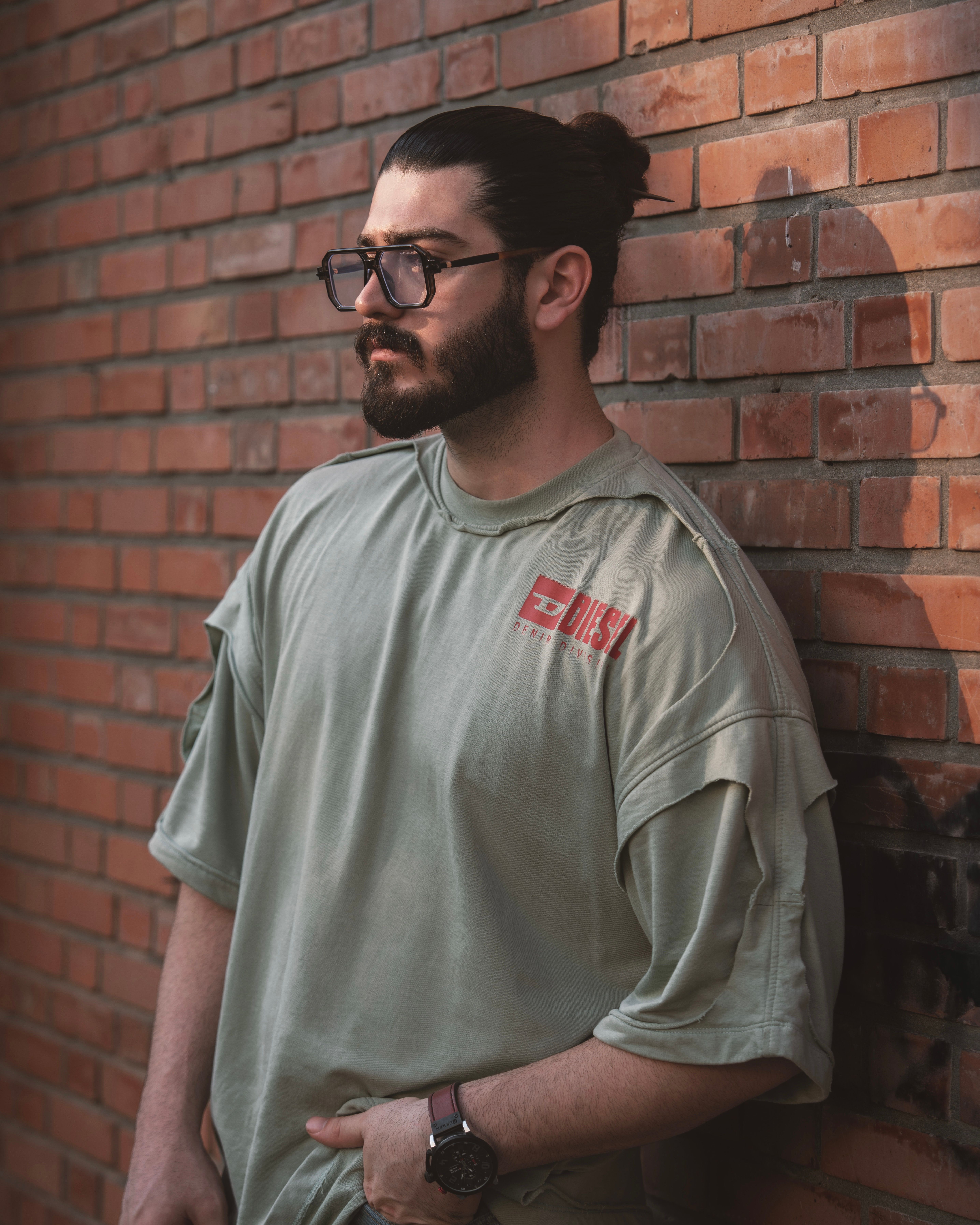 Serious man with beard and glasses leaning against brick wall in urban setting.