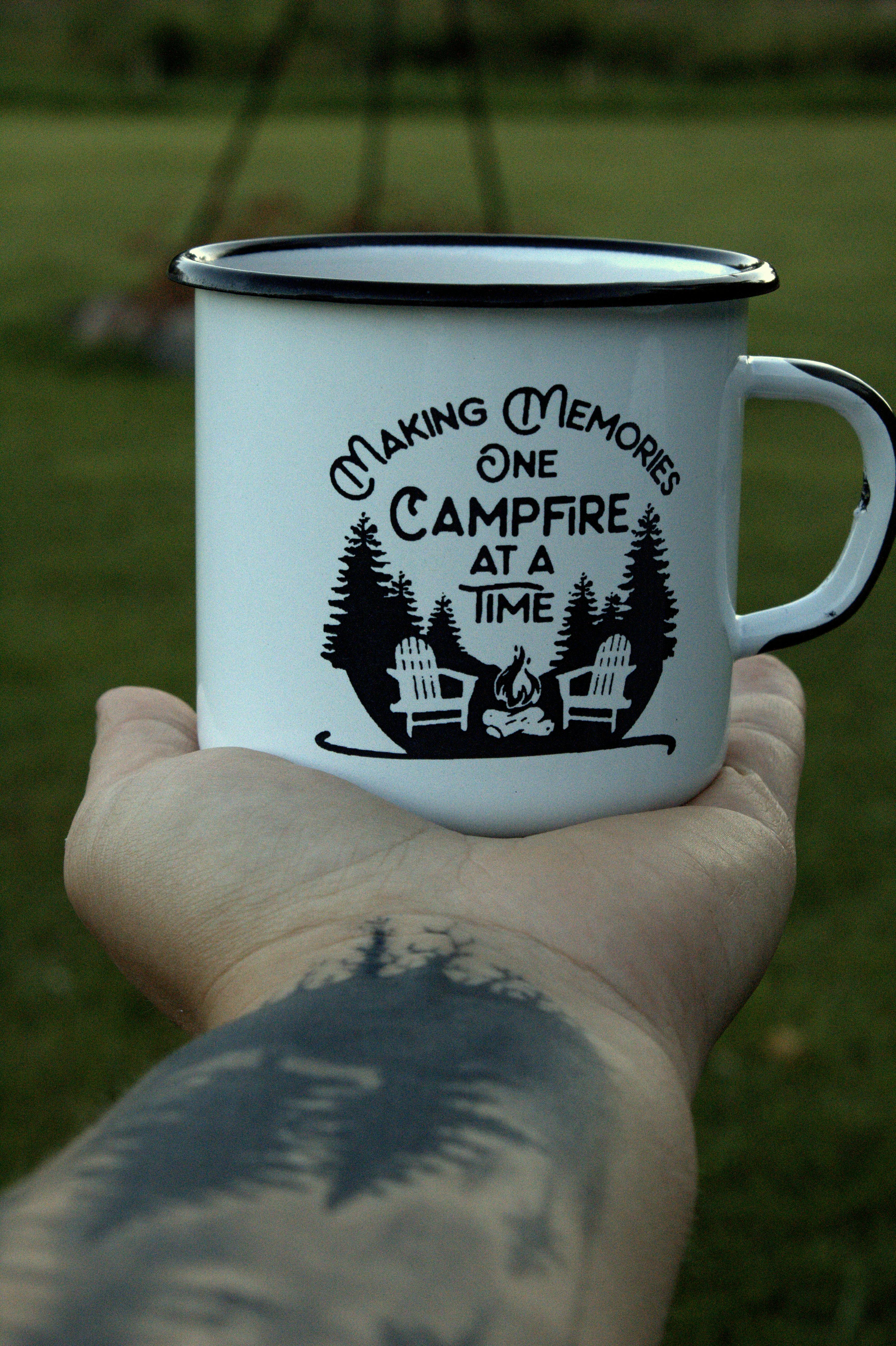 A tattooed hand holds a campfire-themed mug outdoors, perfect for relaxation.