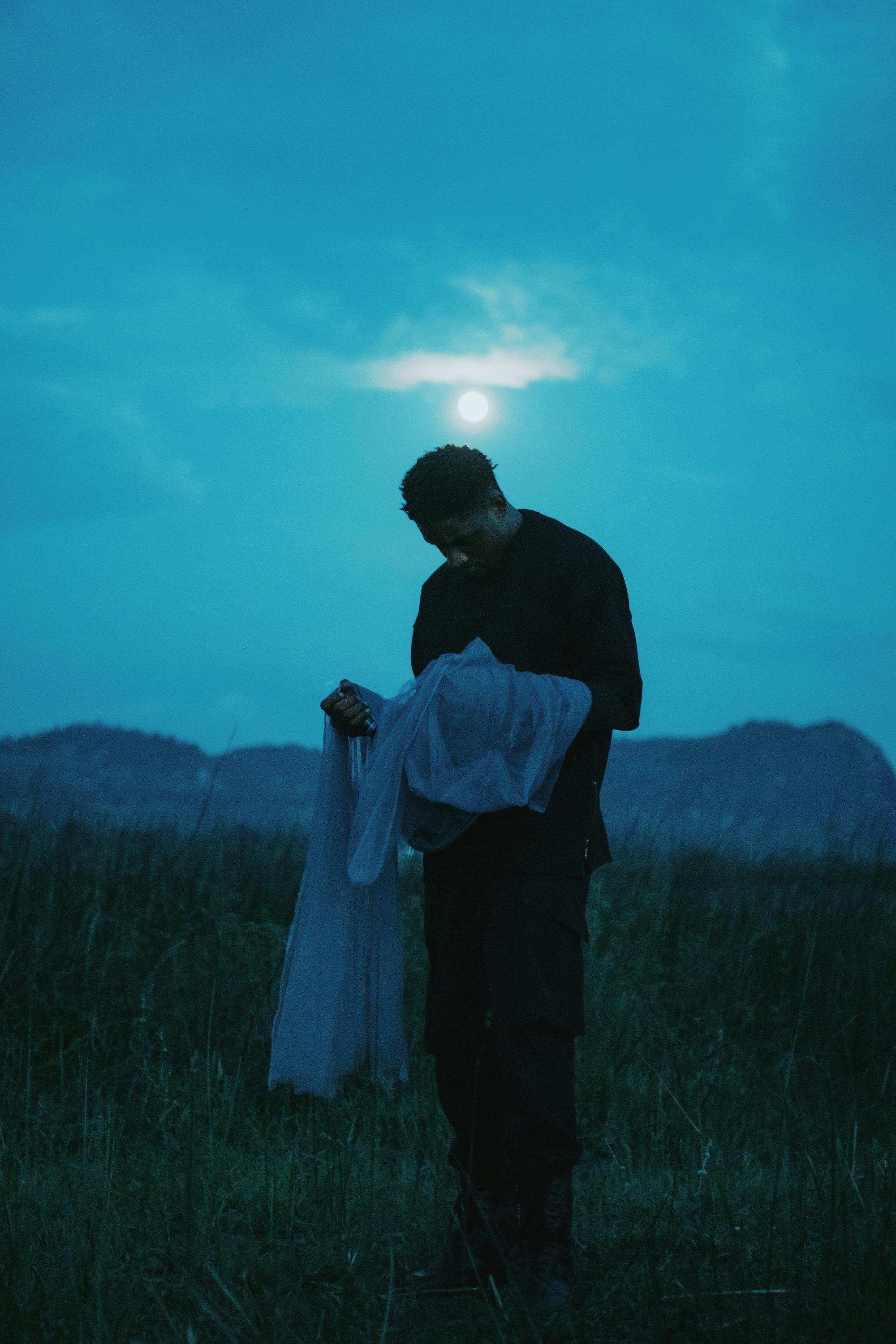 A silhouette of a man holding fabric under a moonlit sky in the Albanian landscape, evoking mystery and serenity.