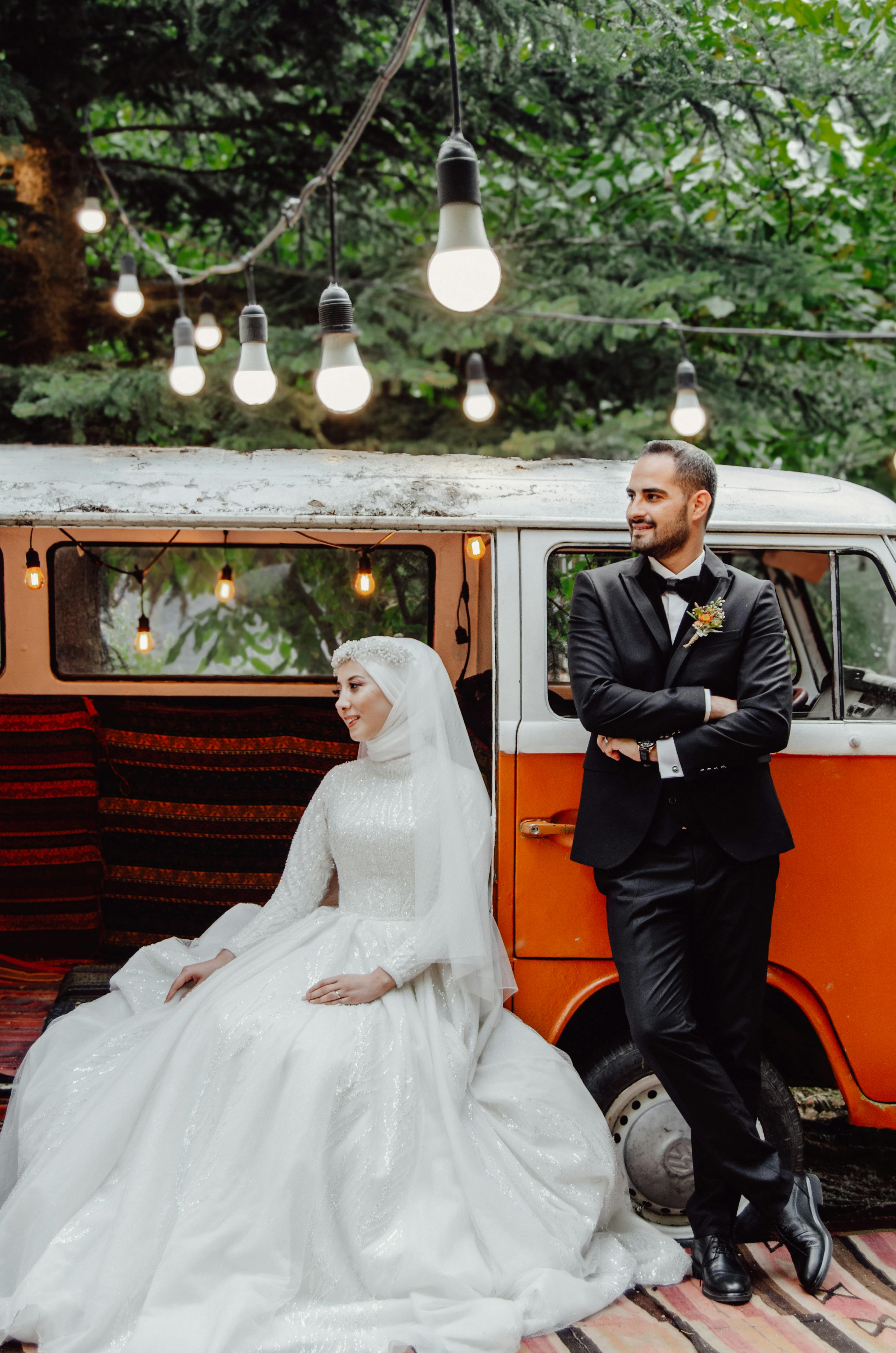 Bride and groom pose beside a vintage orange van adorned with string lights in an outdoor wedding setting.