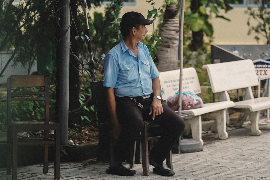 A man in a blue uniform sits on a bench outdoors, surrounded by greenery and urban elements.