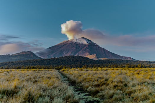A breathtaking view of a volcano emitting smoke during sunrise with a scenic grassy foreground.