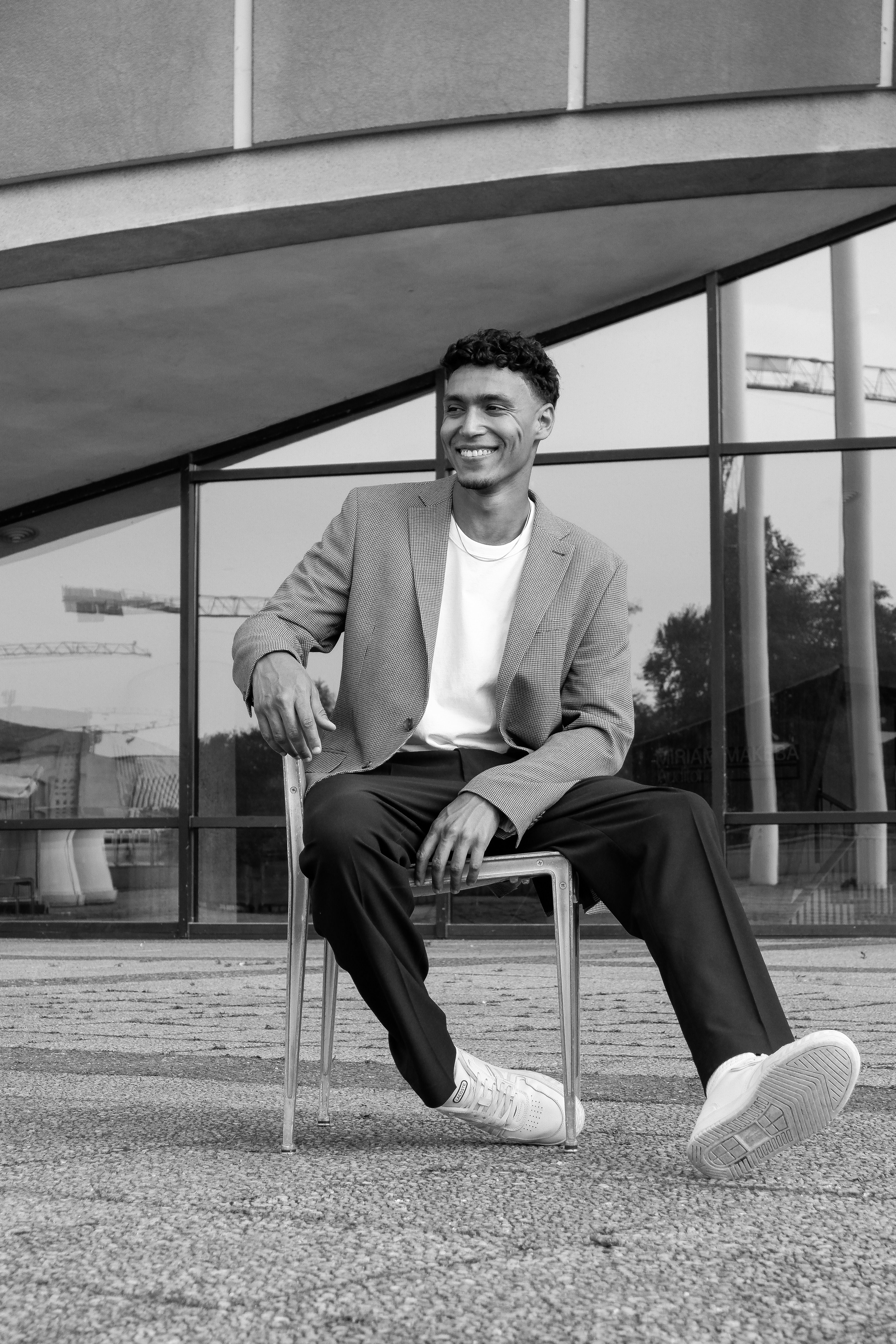 Black and white portrait of a stylish man sitting on a chair in a modern urban environment.