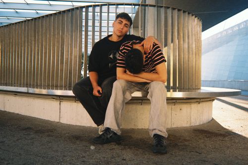 Free Two young men sitting together on a bench in an urban setting, expressing friendship. Stock Photo