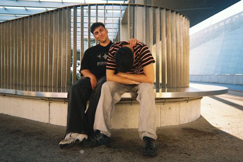 Free Two young men sitting on a modern bench in an urban outdoor setting, interacting casually. Stock Photo