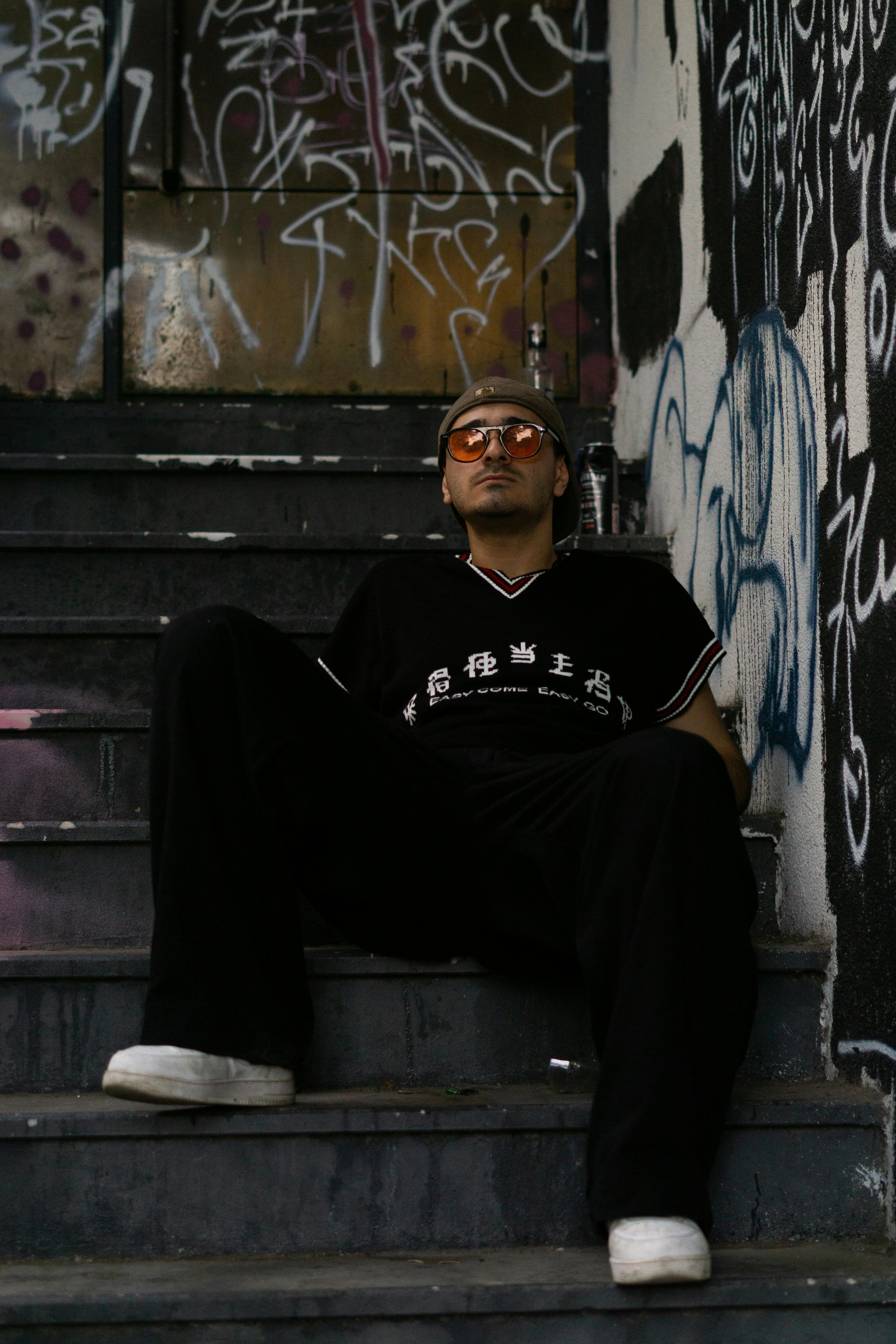 Man in urban attire sitting on stairs with graffiti art, exuding street fashion vibes.