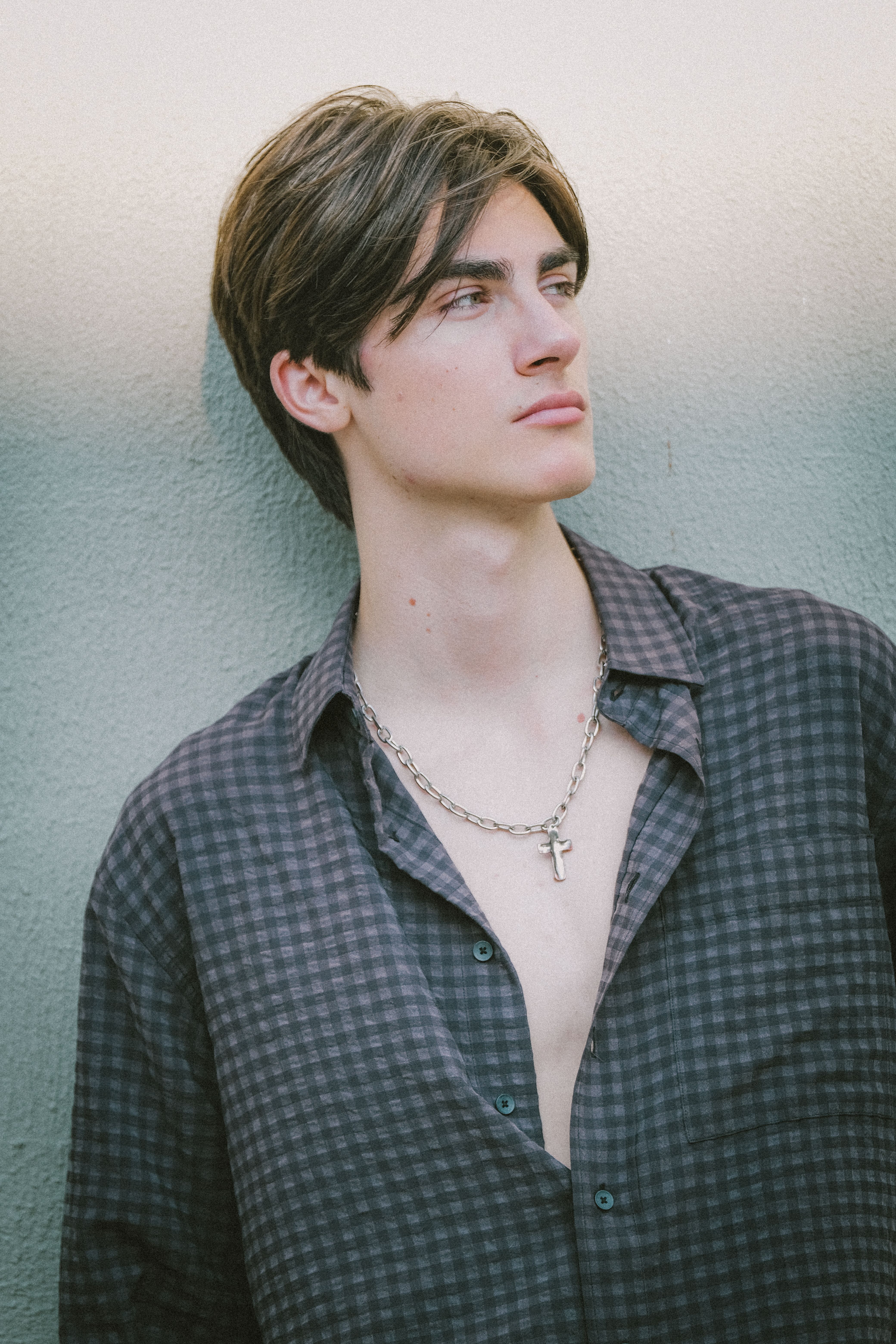 Stylish young man wearing a cross necklace and plaid shirt in a modern urban setting.