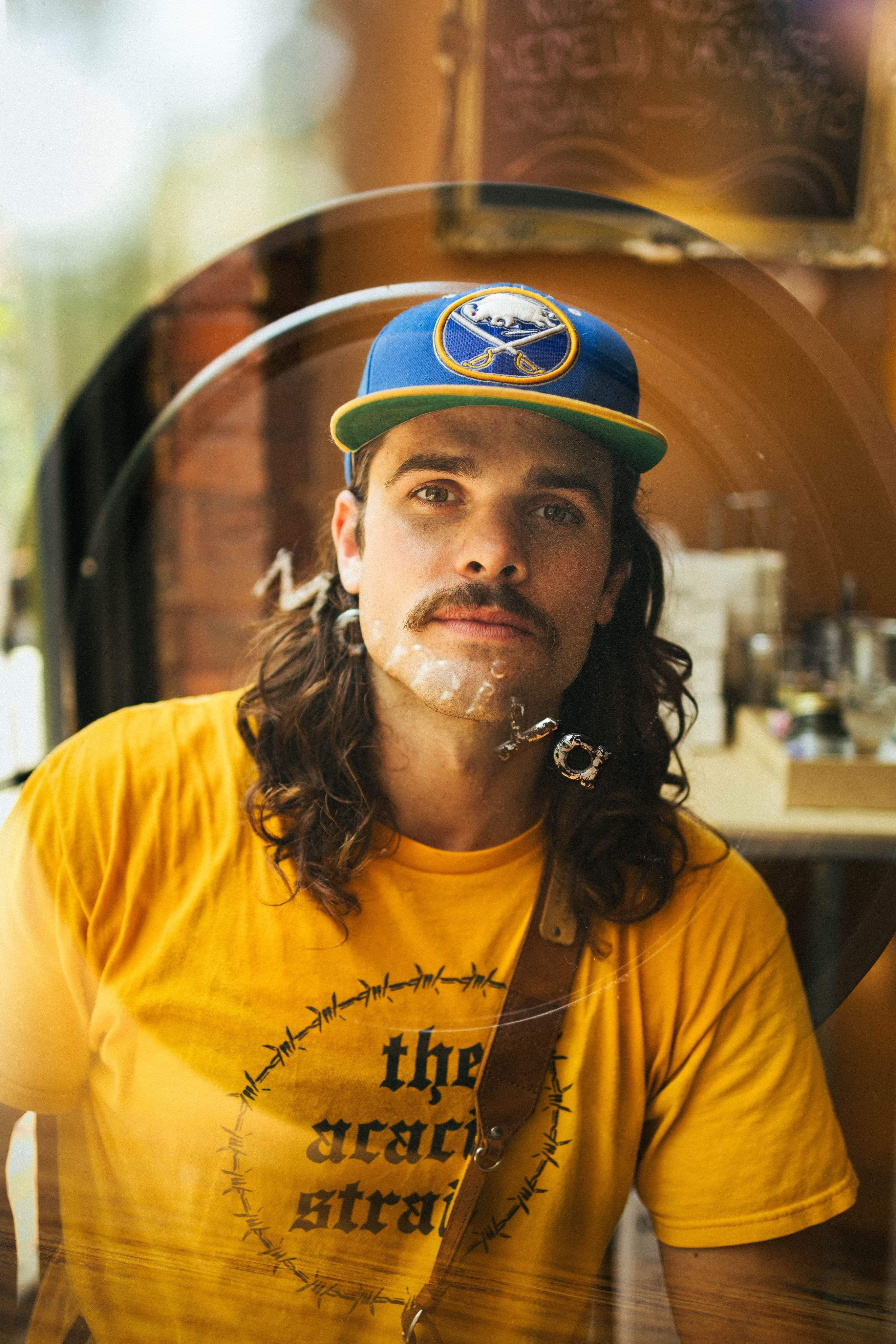 A man with long hair and a mustache wearing a yellow T-shirt and blue cap indoors.
