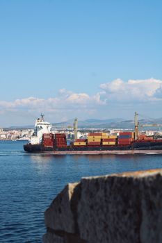 A cargo ship loaded with containers sails past a coastal city with clear blue skies.