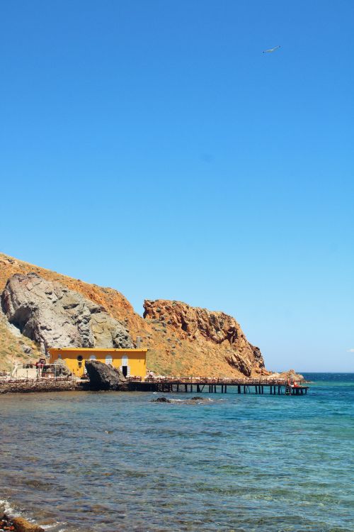 Free A vibrant coastal scene with cliffs, a yellow building, and a wooden pier extending over azure waters under a clear blue sky. Stock Photo