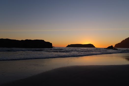Serene sunset over an ocean beach with silhouetted rocks reflecting on the water.