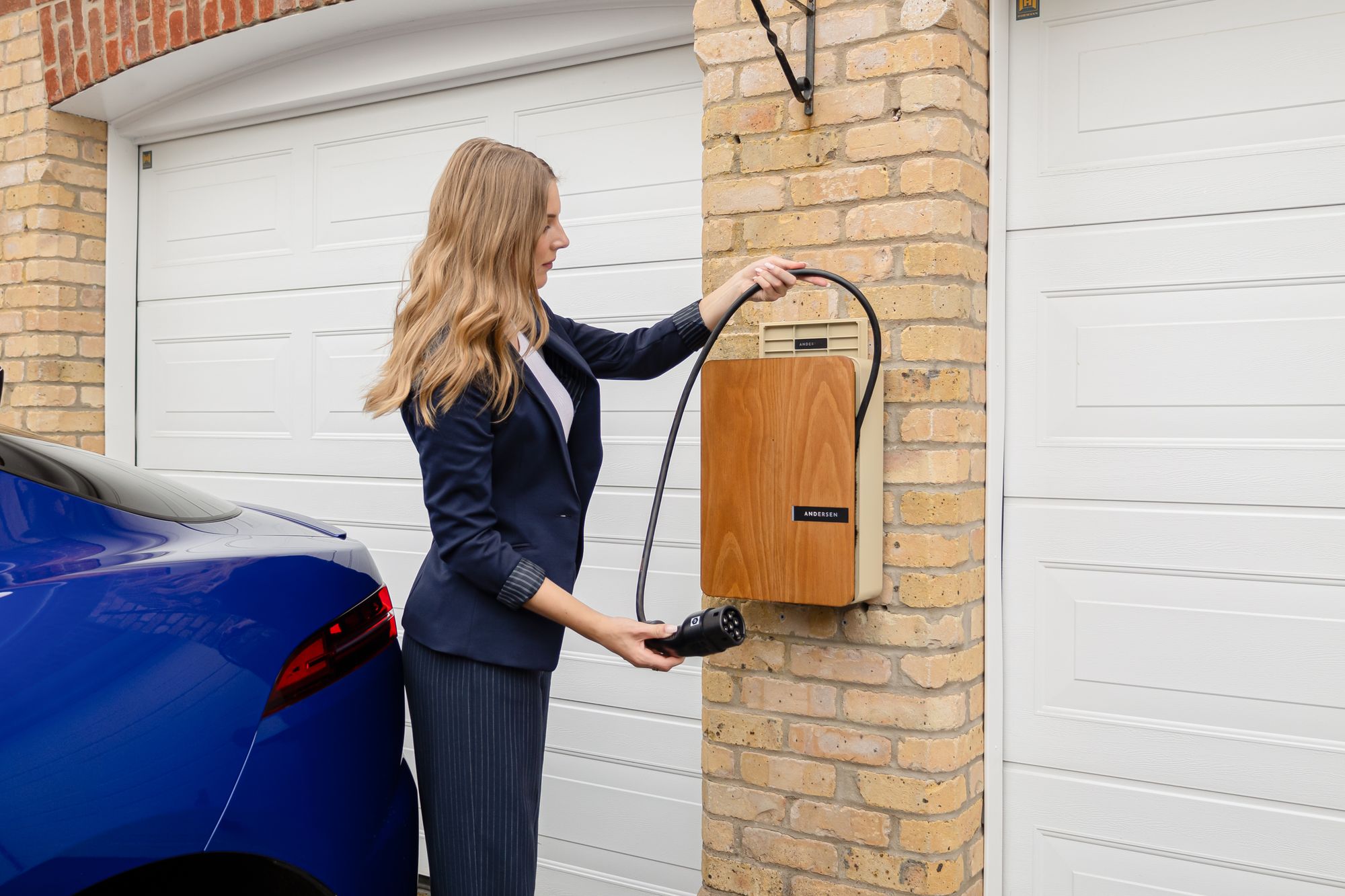 Shows a homeowner using a residential Level 2 wall-mounted EV charger beside a garage, directly illustrating home EV&hellip;