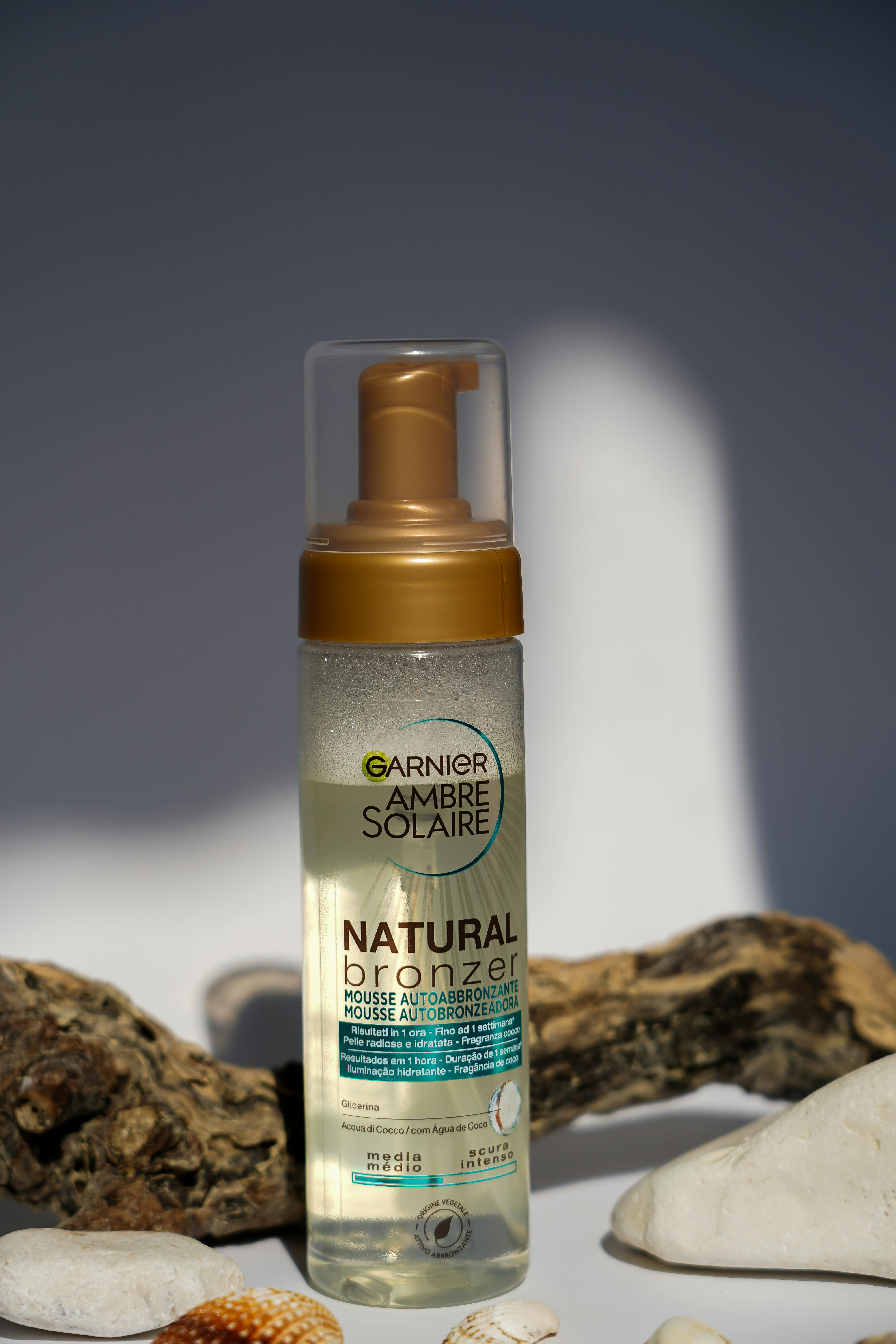 Natural bronzer tanning mousse bottle amidst seashells, photographed in natural light.