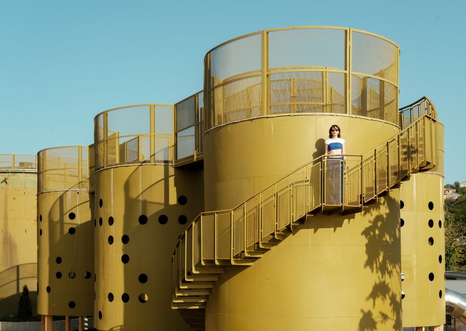 A woman stands on stairs against a yellow silo with a clear blue sky.