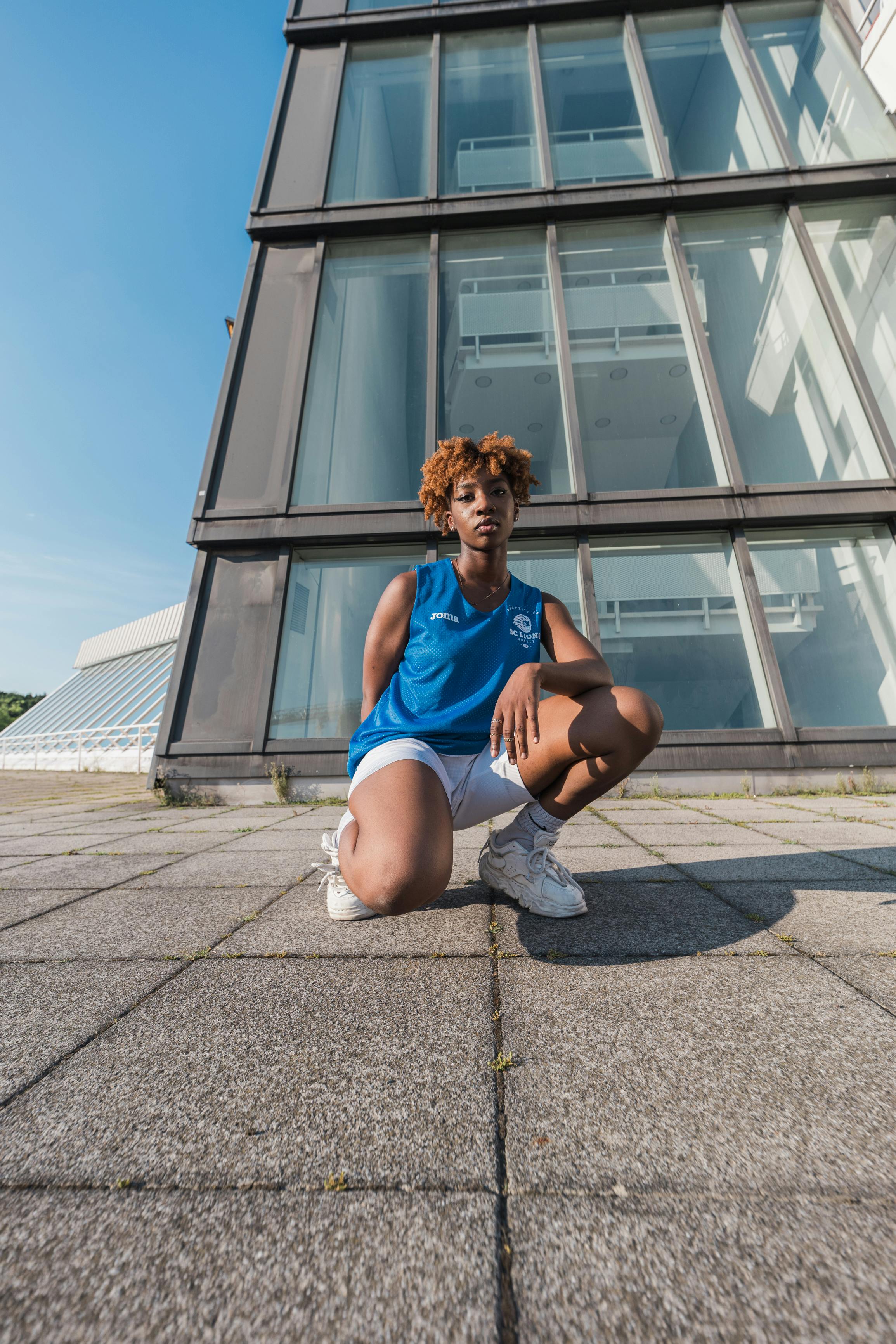 Dynamic urban fashion photo of a woman crouching in front of a Berlin building with large windows, showcasing sportswear.