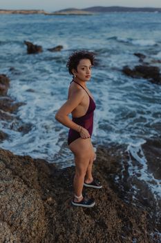A woman in a swimsuit stands on a rocky beach during sunset, watching the waves crash.