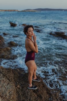A woman in a purple swimsuit stands on rocks by the ocean at sunset, embracing nature's beauty.