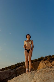 A woman in a bikini stands on a rocky beach, gazing into the distance at sunset.