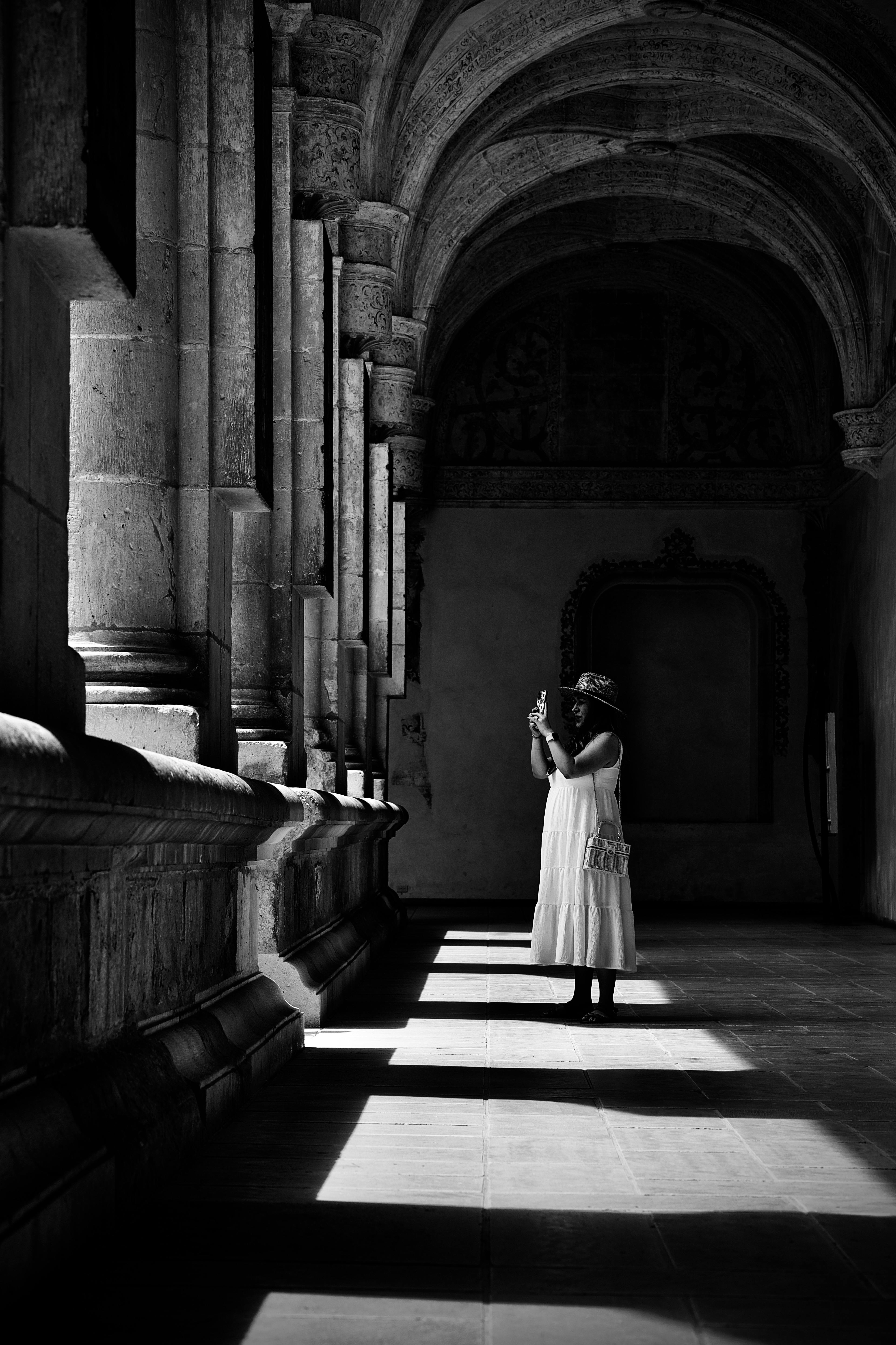 Black and white image of woman in sun hat exploring monastery corridor with dramatic light and shadows.