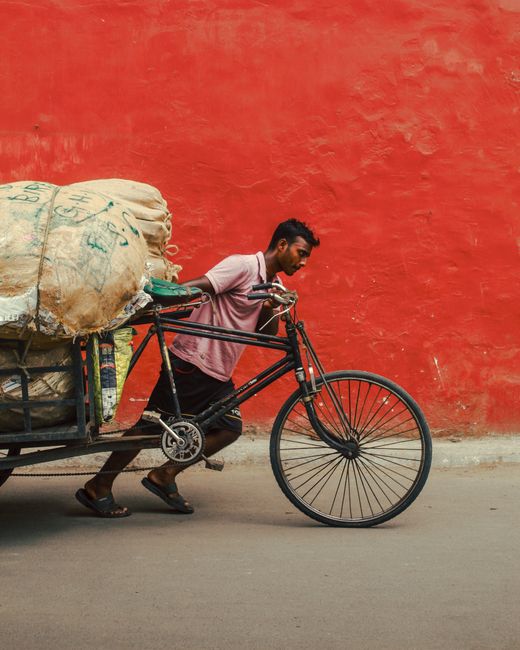 Man exerting effort to push a loaded cart on the street, vibrant red wall backdrop.
