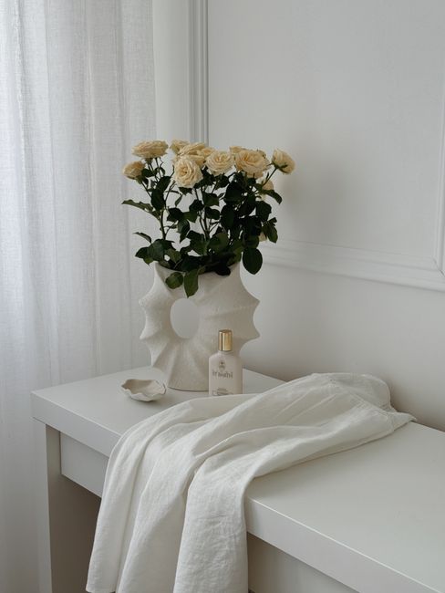 Aesthetic arrangement of cream roses in a white vase on a minimalist dresser with soft fabric.