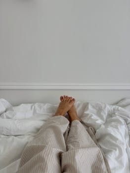 Relaxing morning in bed with legs covered in pajamas on white sheets. Tranquil indoor setting.