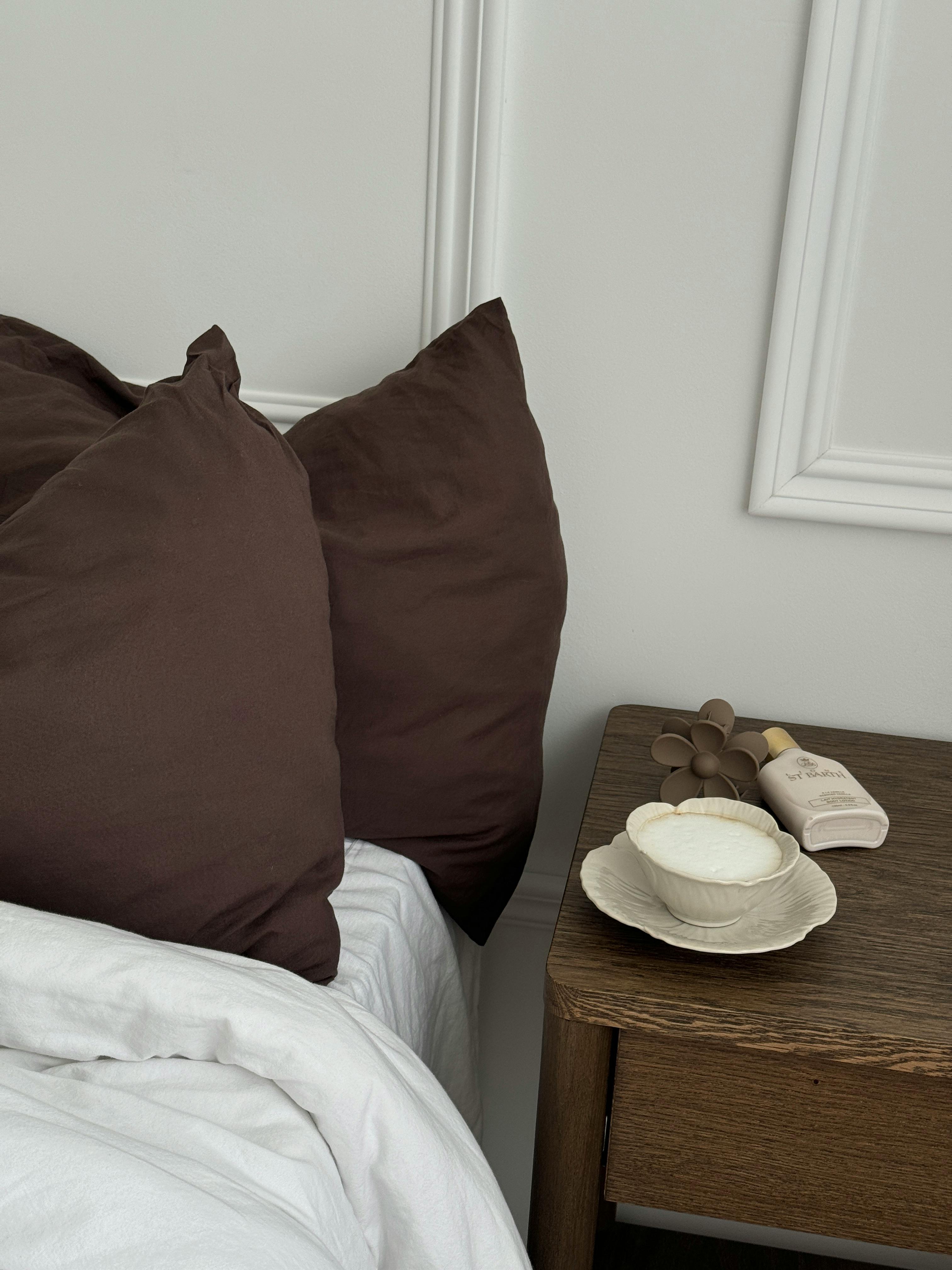 A cozy bedroom scene featuring brown pillows, a nightstand with a bowl, and skincare items.