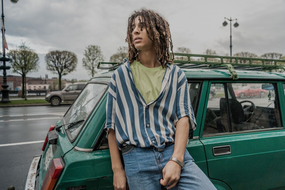 A stylish young man with dreadlocks poses by a vintage car in a city setting.