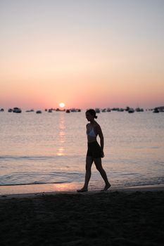 Silhouetted woman strolling on a beach with a serene sunset backdrop, evoking relaxation and leisure.