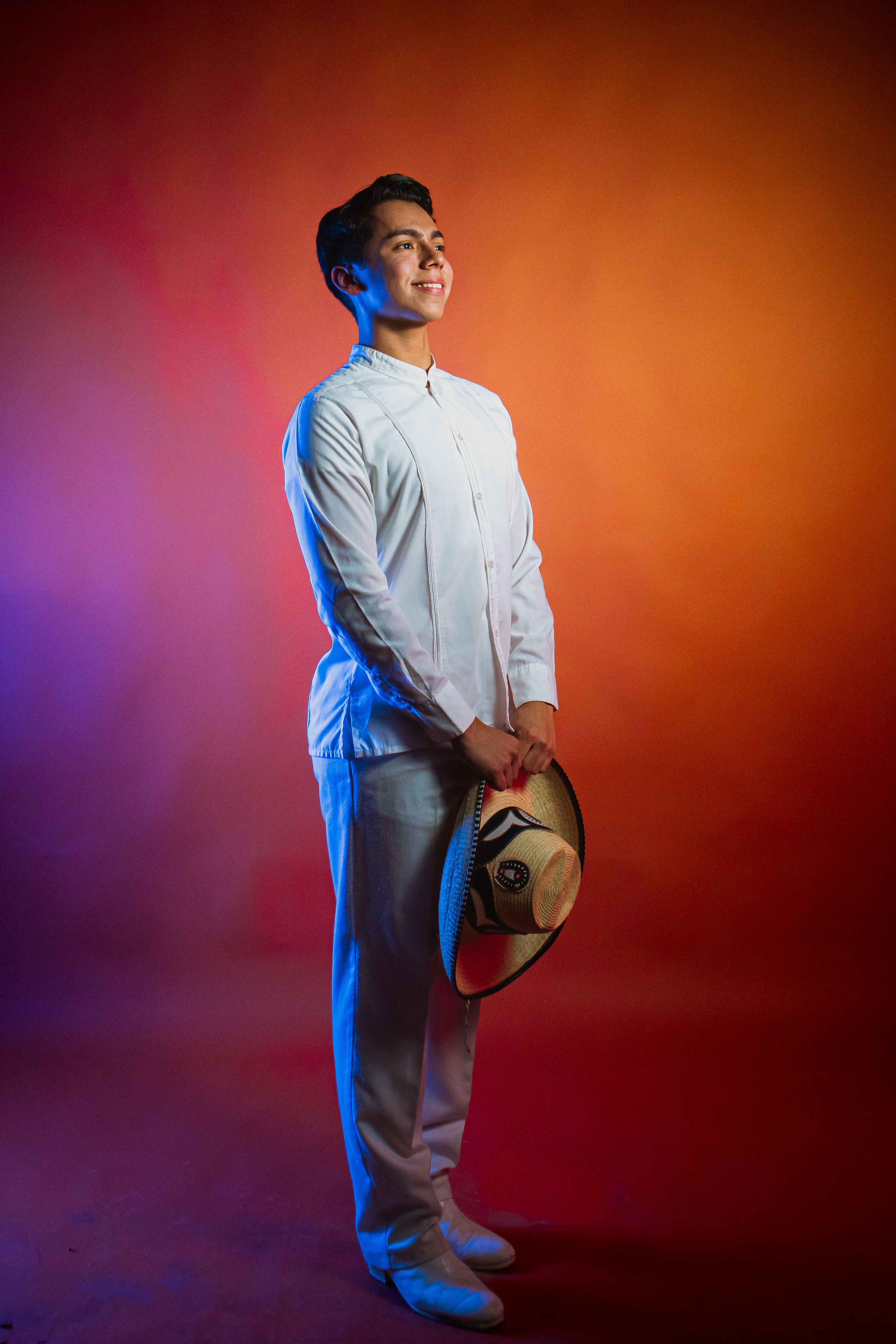 Portrait of a young man in traditional Mexican attire holding a sombrero, against a colorful gradient background.