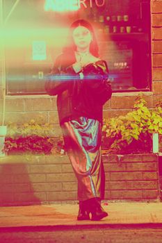 A woman in fashionable attire stands illuminated by vibrant city lights, creating a retro ambiance.