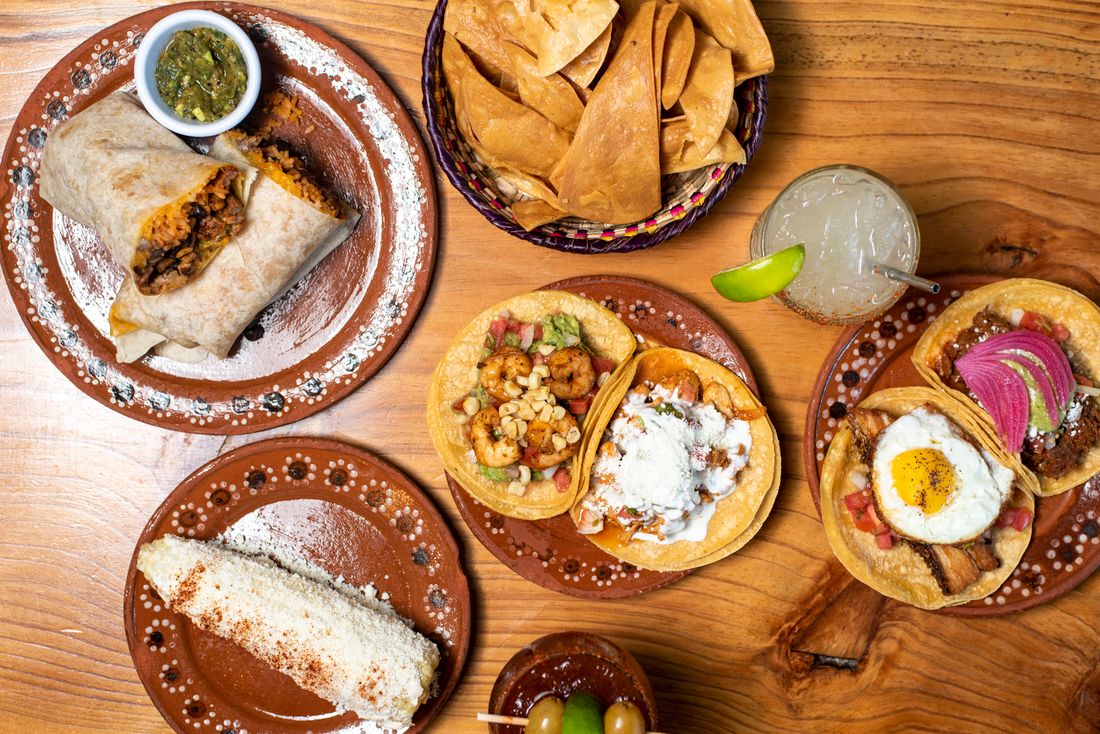 Overhead spread of burrito, tacos, fried egg taco, street corn, chips and a lime drink
