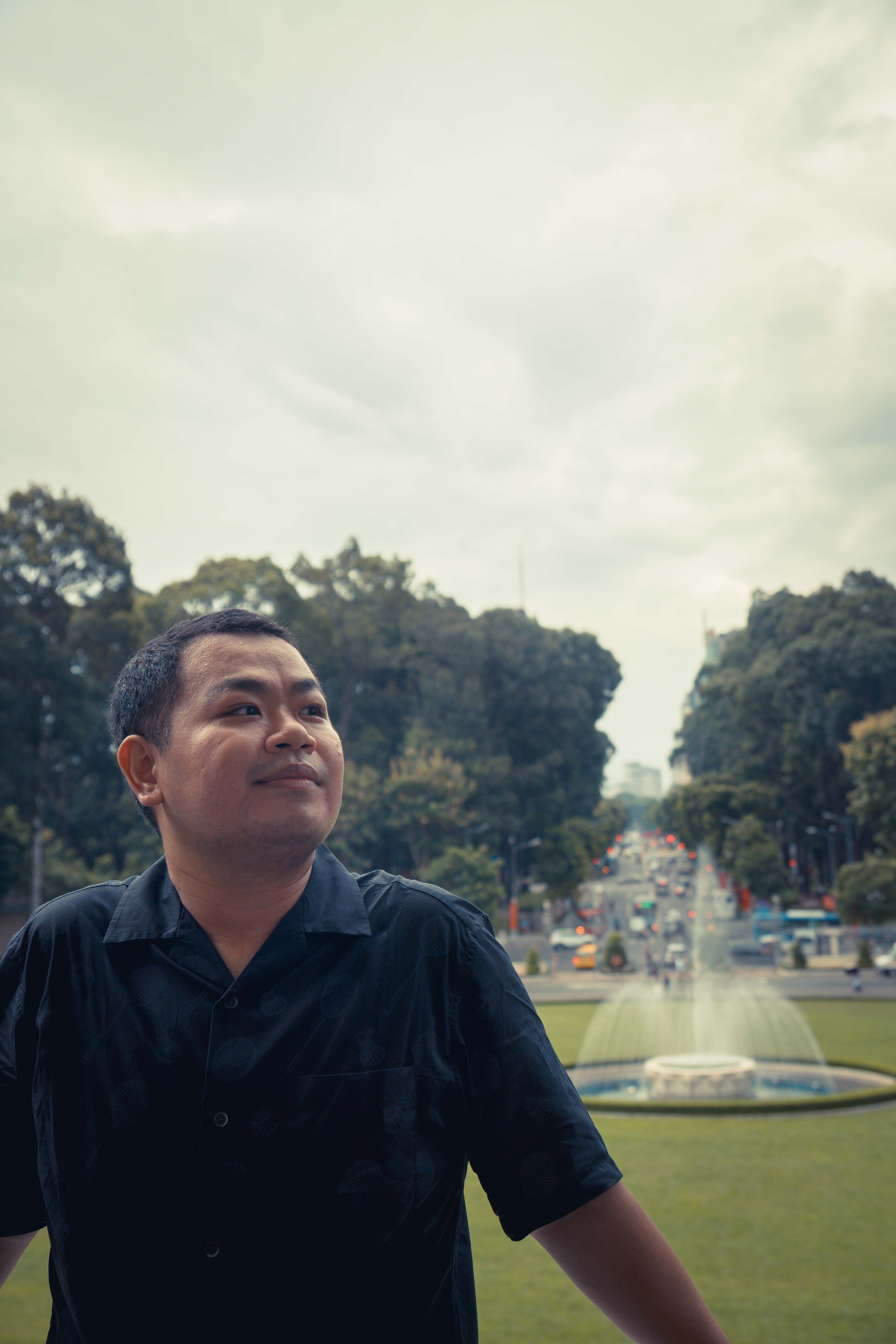 A man standing near a fountain in Ho Chi Minh City park with lush greenery and city backdrop.