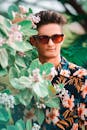 一位时尚的年轻人戴着墨镜、穿着花卉衬衫，在郁郁葱葱的绿叶和花蕾间摆出姿势