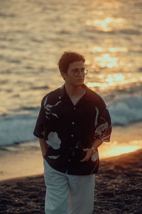 Free A man walks along the beach with a camera during sunset, wearing a patterned shirt and glasses. Stock Photo