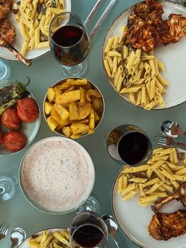 Top view of a Mediterranean meal with pasta, grilled meats, and red wine on a table.