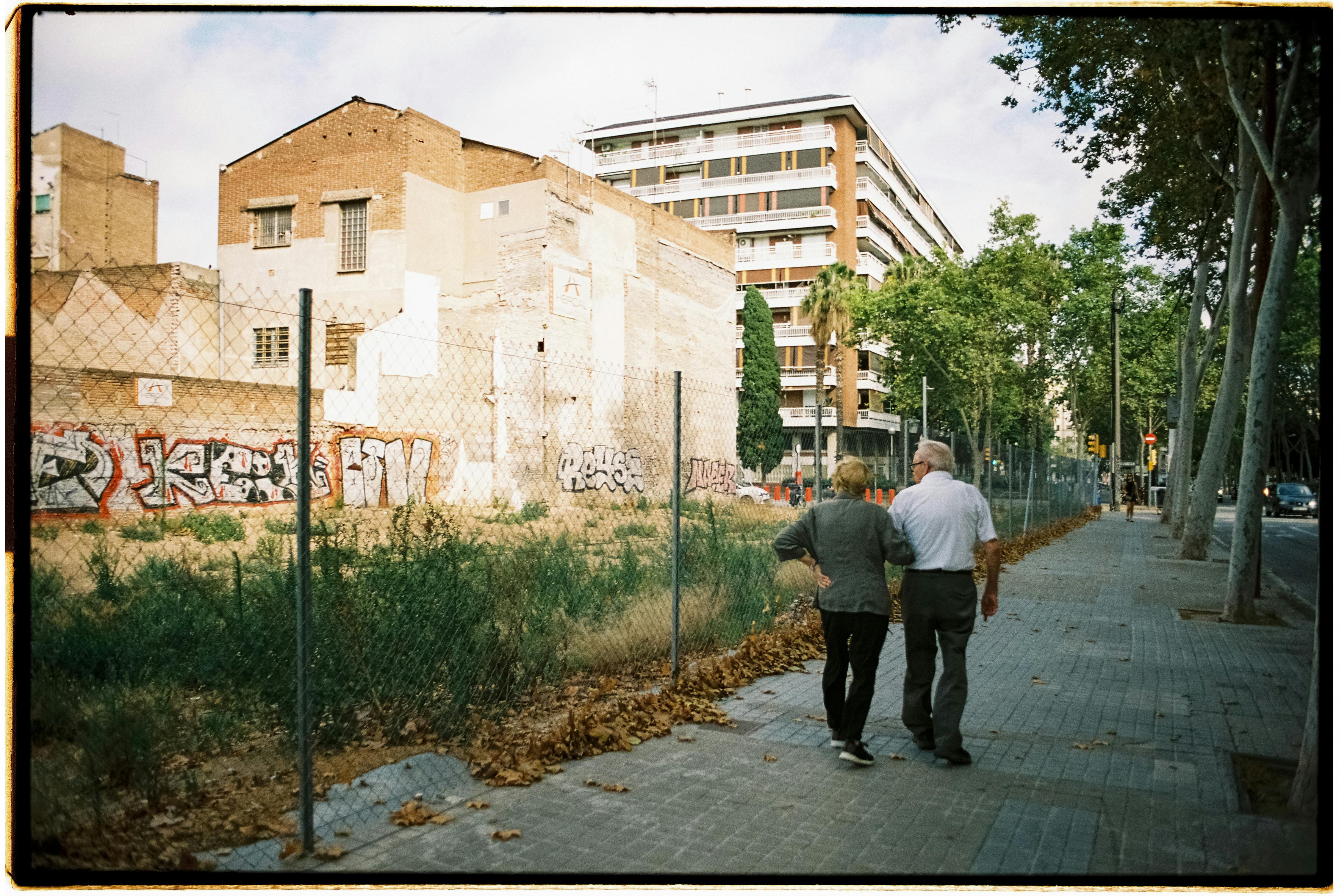 An elderly couple strolls down a city sidewalk, surrounded by urban buildings and graffiti.