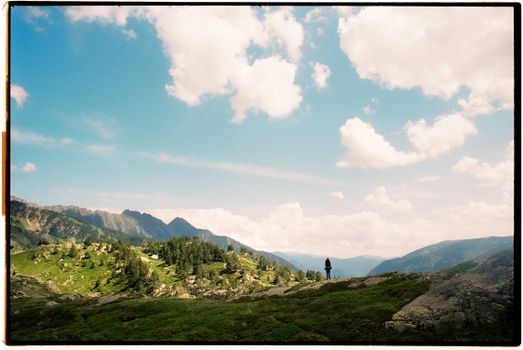 A lone hiker stands in a lush, mountainous landscape under a vibrant blue sky with clouds.