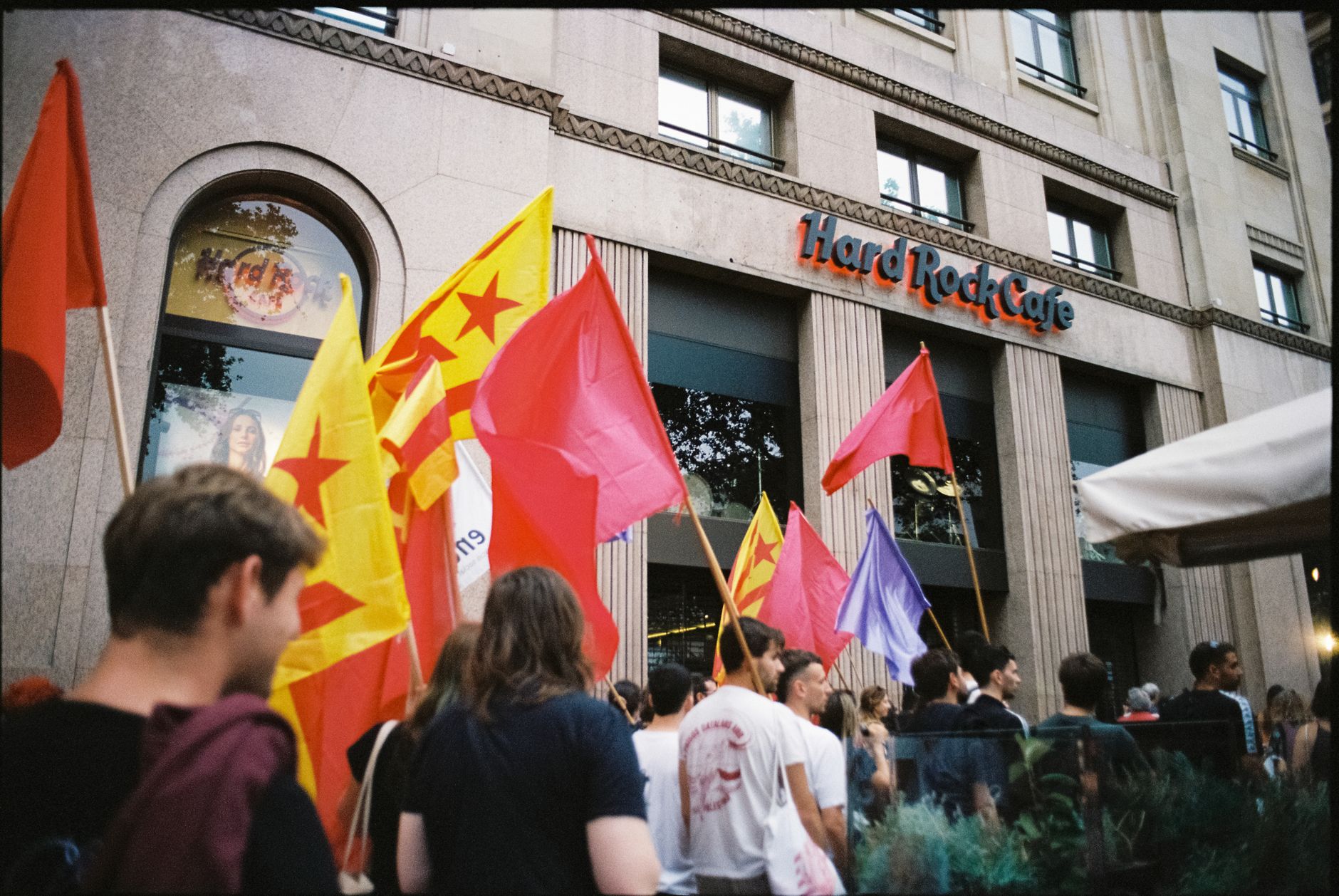 A group of protesters waving colorful flags outside the Hard Rock Cafe on a city street in daylight.