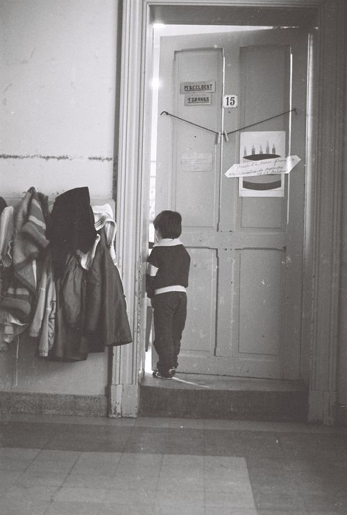 Free Black and white image of a child peeking into a classroom, coats hanging nearby. Stock Photo