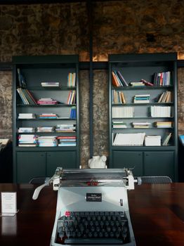 A classic typewriter on a wooden desk in a library with bookshelves and stone walls.