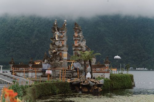 Free A scenic view of a Balinese temple surrounded by water and mountains under a misty sky. Stock Photo