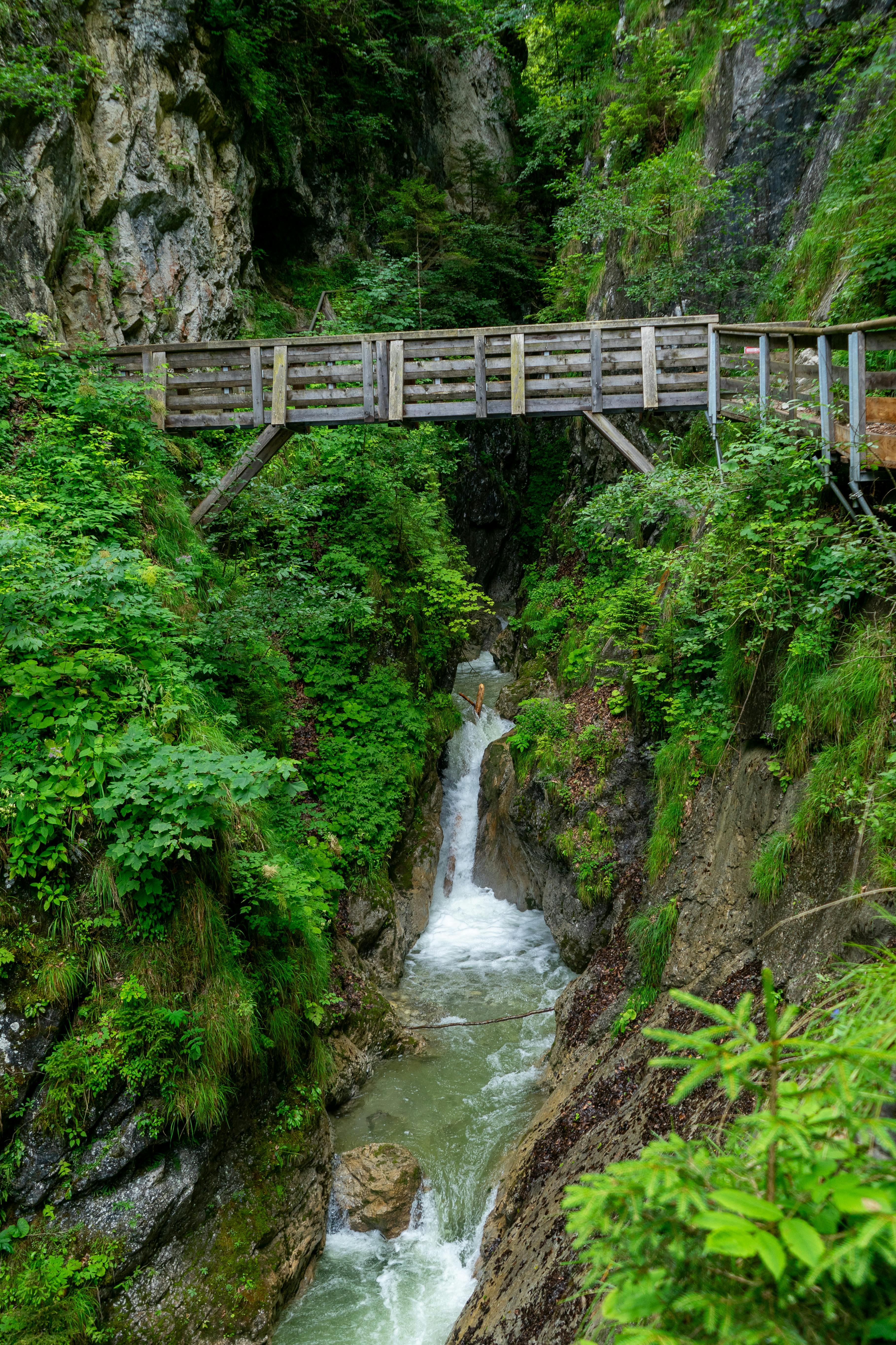 A picturesque wooden bridge spans a lush green gorge with a cascading waterfall in Stans, Tirol.