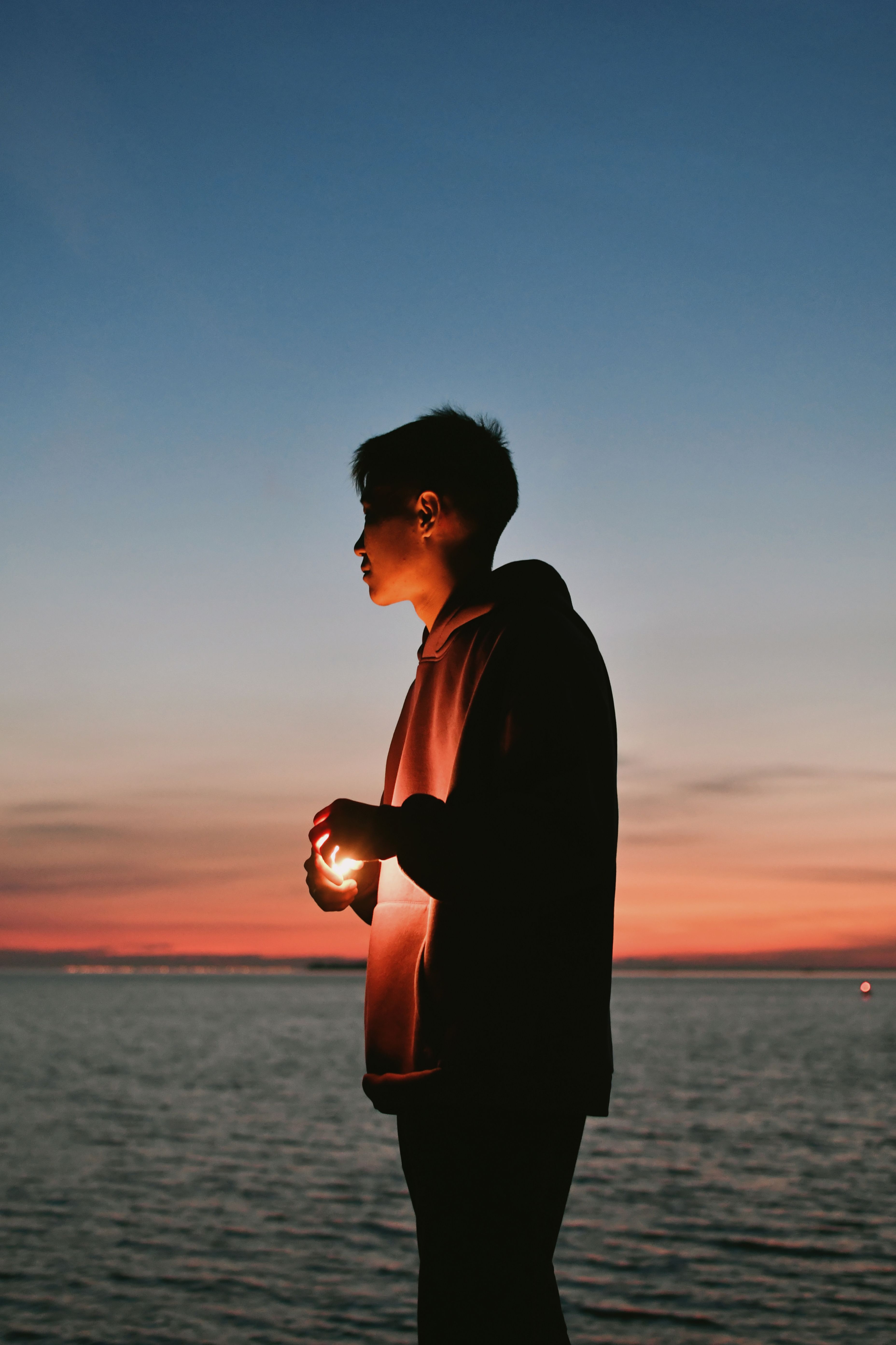 A solitary figure holding a glowing light stands against a sunset over the calm sea.