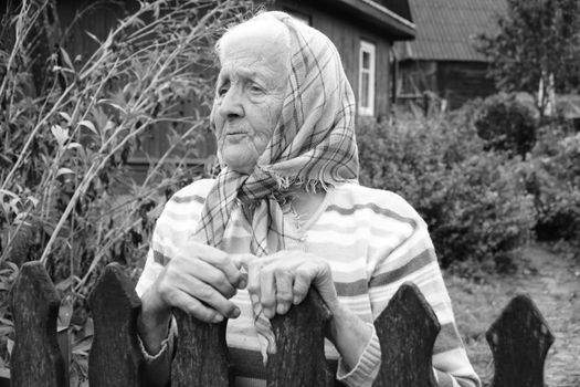 Black and white portrait of an elderly woman with a headscarf standing by a wooden fence.