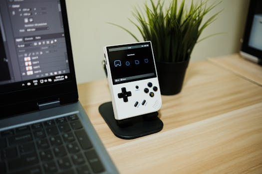 A handheld gaming console on a wooden desk next to a laptop and plant.
