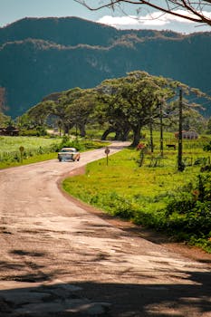 A classic car drives along a picturesque road in Cuba, surrounded by lush greenery and mountains.